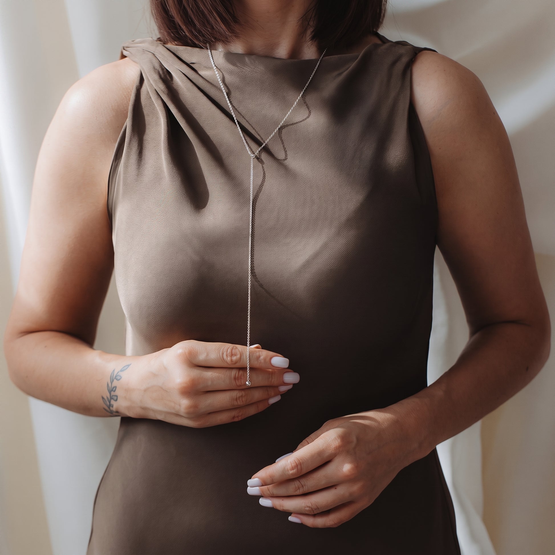 Gros plan d’un mannequin portant un collier lariat bola en argent 925 sur une robe taupe sans manches, tenant délicatement la chaîne pendante.