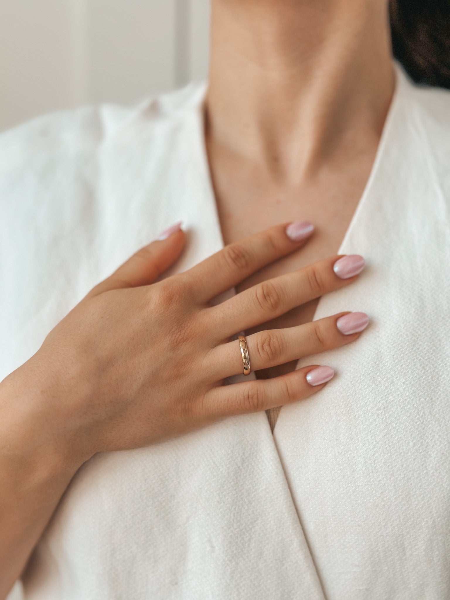 Gros plan d'une main avec une manucure rose reposant sur la poitrine, portant une alliance en or et un vêtement texturé blanc.