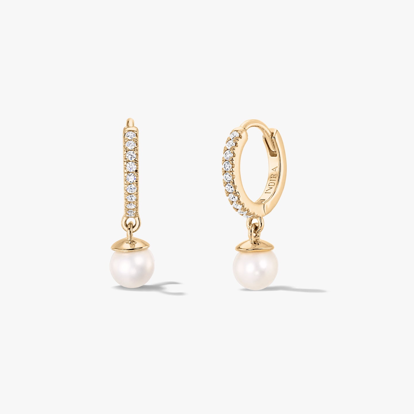 Pair of gold‑tone silver huggie hoop earrings with pavé zirconia and white natural pearl drops by Indira, shown front and side on a white background.