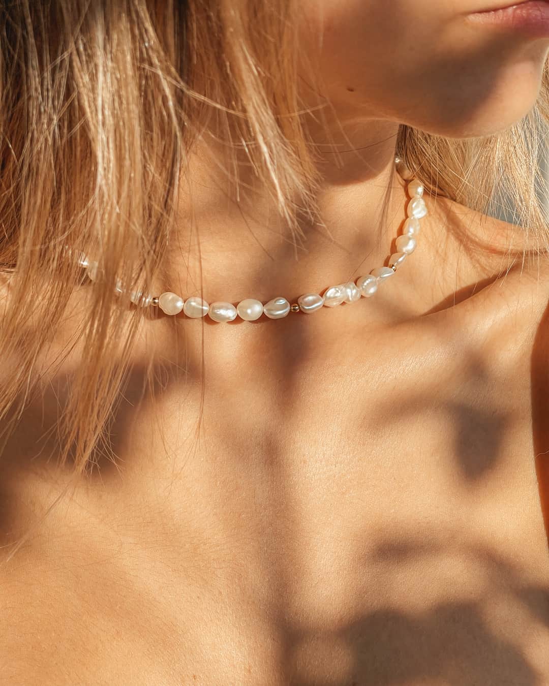 Gros plan d'une femme portant un collier en argent et perles avec des accents dorés, la lumière du soleil mettant en valeur le teint et la texture de la peau.