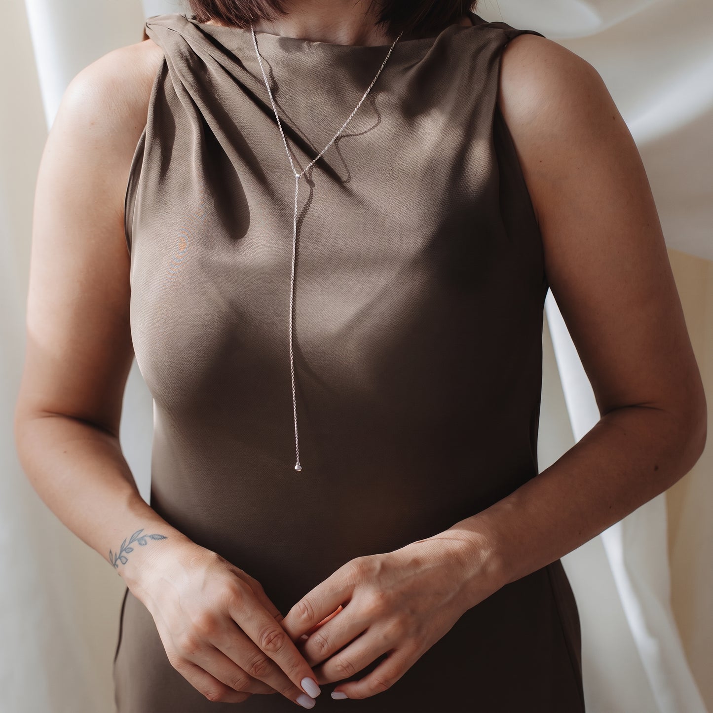 Gros plan d’une personne portant un collier bola (lariat) en argent sterling sur une robe en satin taupe, dans une douce lumière intérieure.
