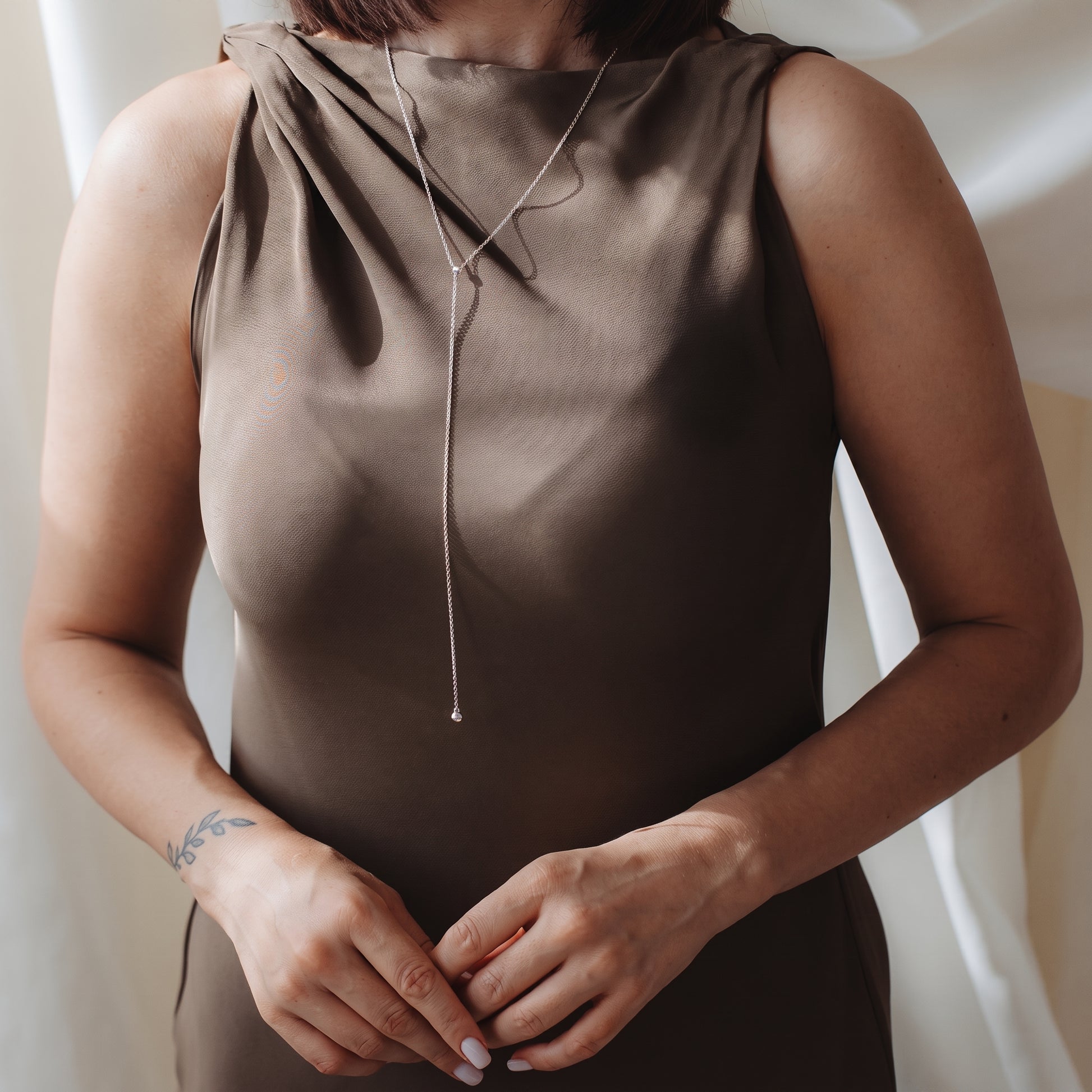 Gros plan d’une personne portant un collier bola (lariat) en argent sterling sur une robe en satin taupe, dans une douce lumière intérieure.