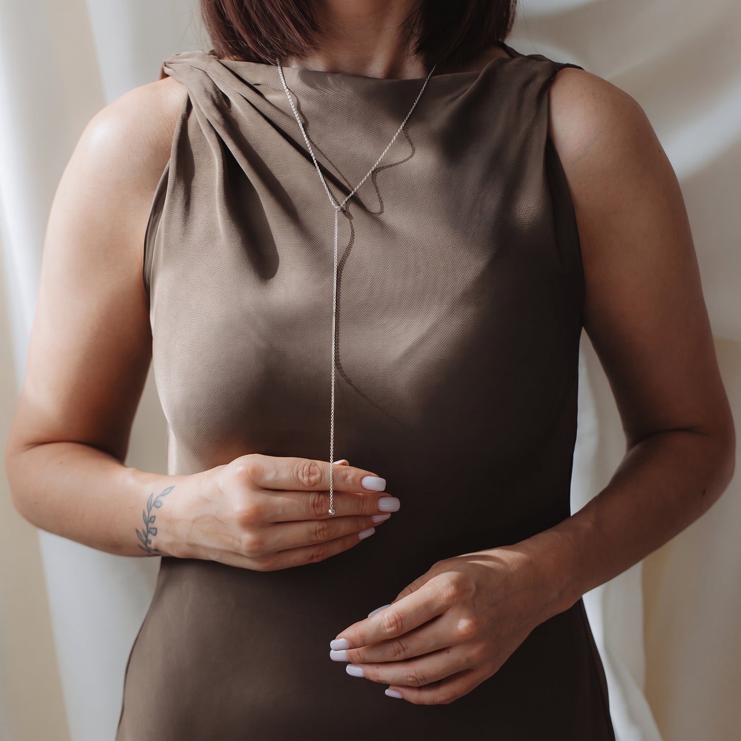 Gros plan d’un mannequin portant un collier lariat bola en argent 925 sur une robe taupe sans manches, tenant délicatement la chaîne pendante.