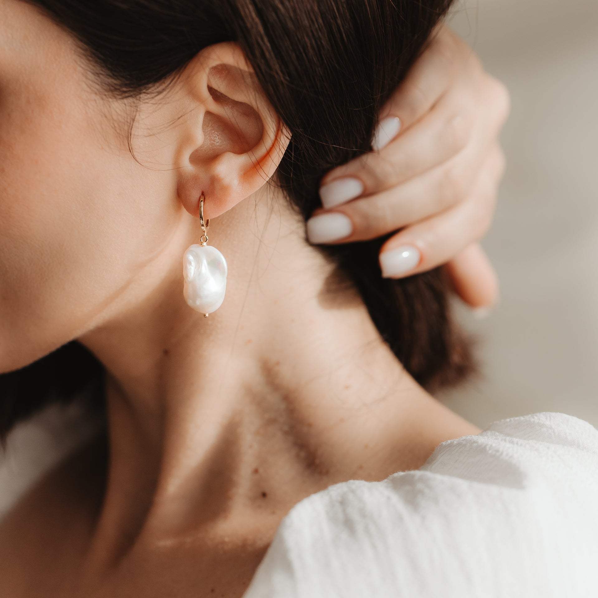 Gros plan d’une oreille portant une créole en argent 925 plaqué or avec une grande perle baroque blanche ; une main à la manucure neutre près de l’oreille sous une douce lumière naturelle.