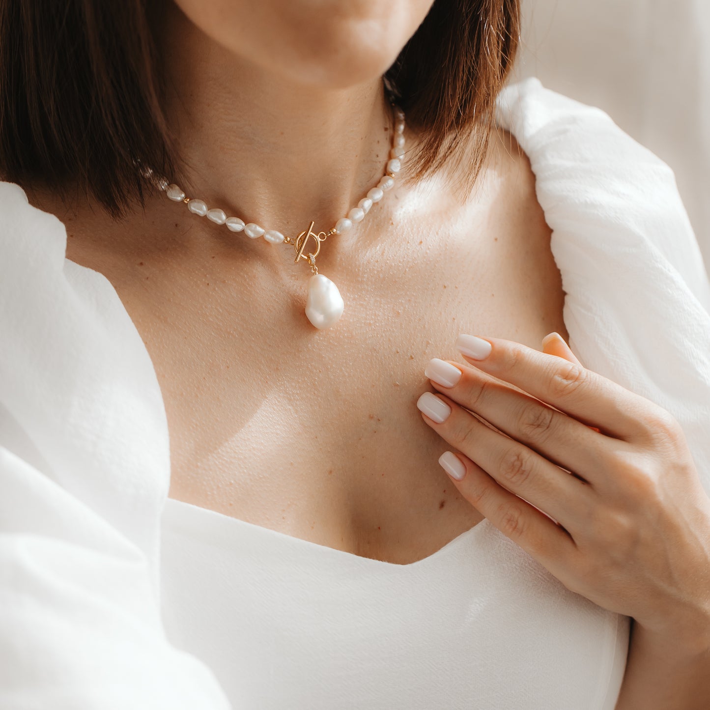 Gros plan d’une femme portant un rang de perles naturelles avec un pendentif en perle baroque plaqué or, touchant sa clavicule sous une douce lumière naturelle.