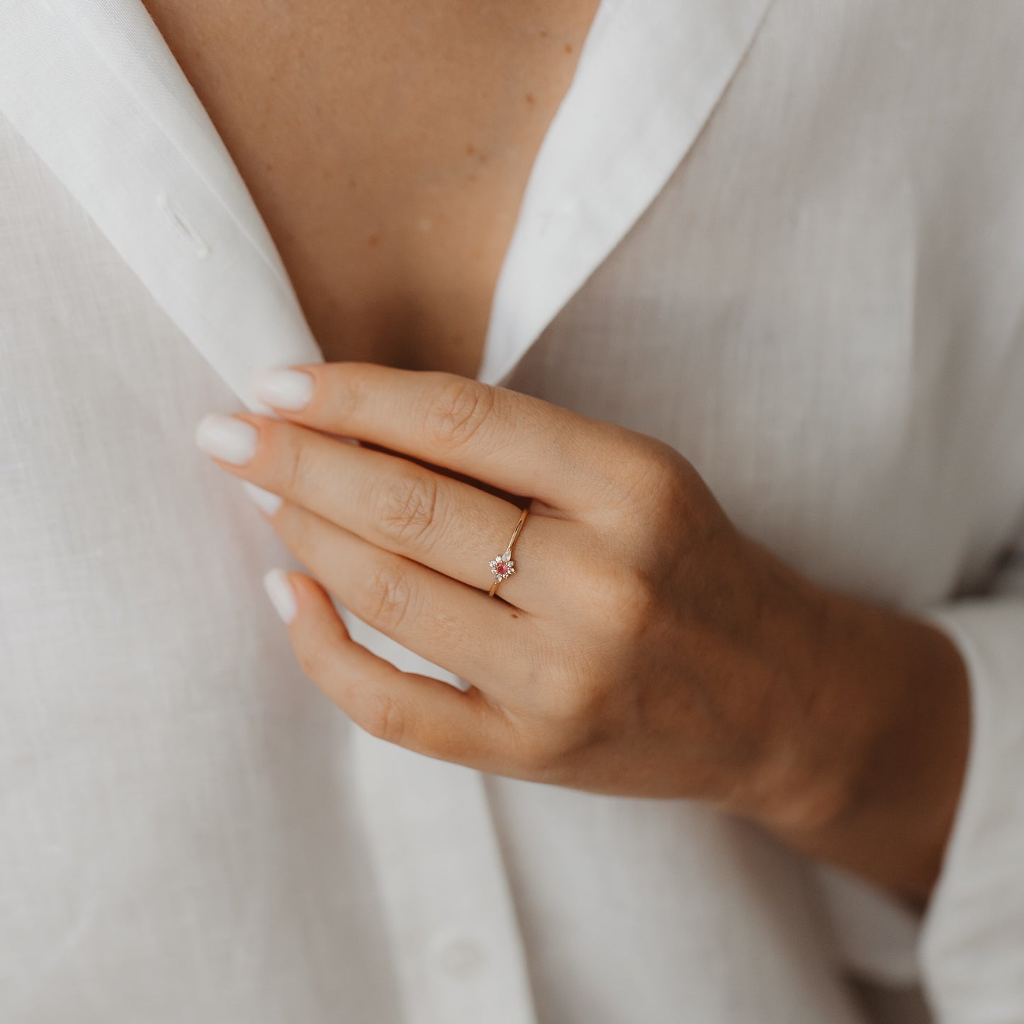 Gros plan d’une main portant la bague Flora en or jaune 14K, sertie d’un saphir blanc et d’un motif floral en tourmaline rose, contre une chemise en lin blanche.