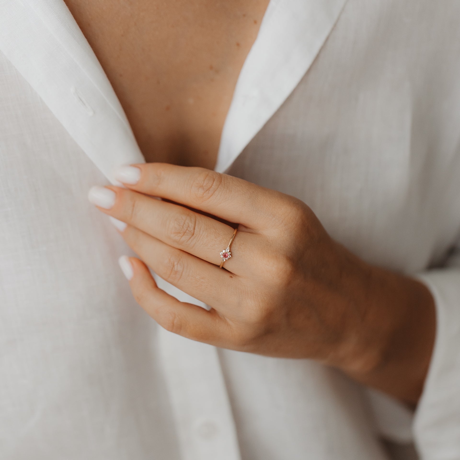 Gros plan d’une main portant la bague Flora en or jaune 14K, sertie d’un saphir blanc et d’un motif floral en tourmaline rose, contre une chemise en lin blanche.