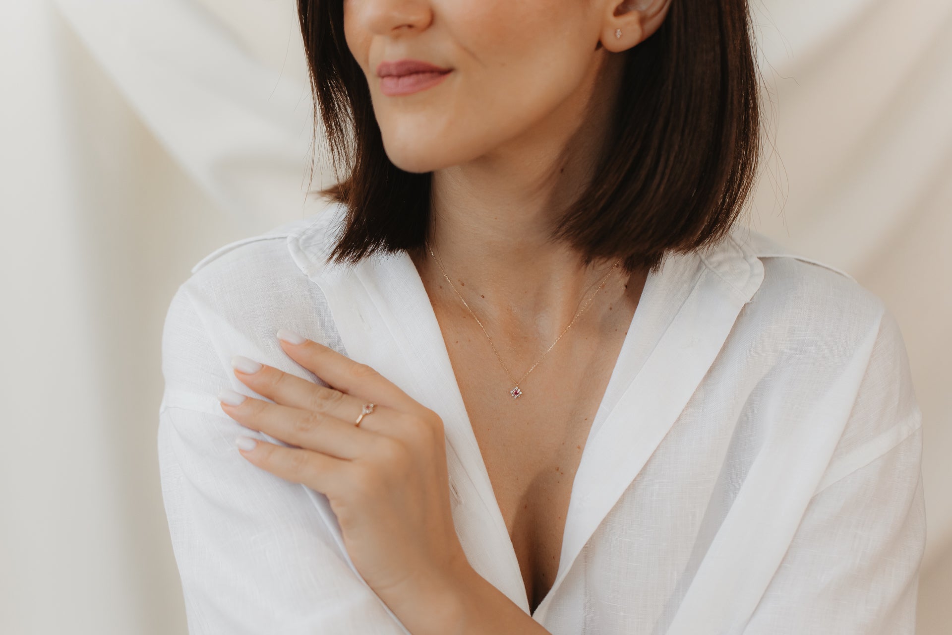Gros plan d'une personne en chemisier blanc portant une bague délicate en or jaune 14K sertie d'une tourmaline rose et d'un saphir blanc, la main posée sur l'épaule sur un fond doux et neutre.