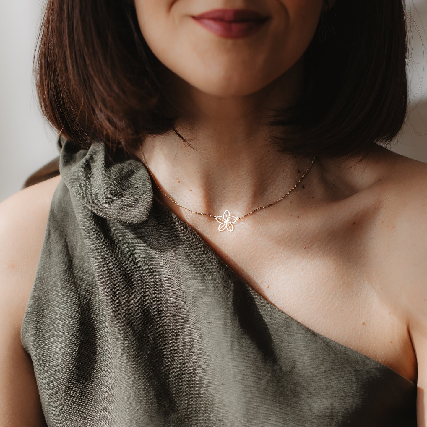 Close-up of a person wearing a delicate 14K yellow gold wildflower outline necklace at the collarbone, styled with an olive one-shoulder top in natural light.