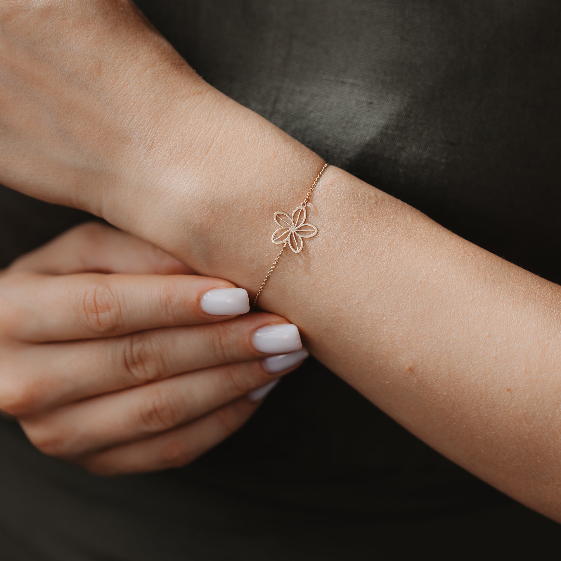Close-up of a delicate 14K gold bracelet featuring an openwork wildflower charm on a wrist, against a dark fabric background.