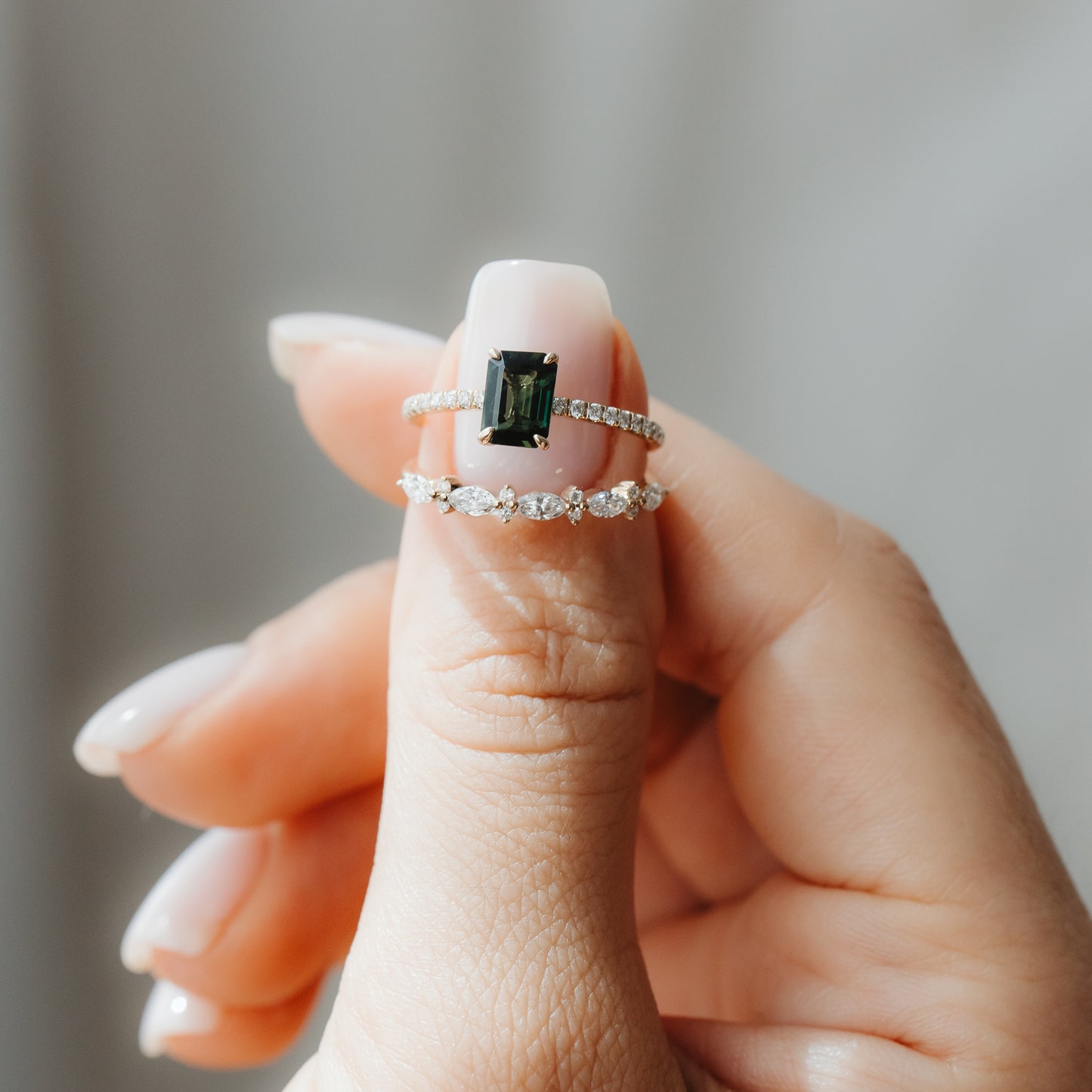 Close-up of a hand holding two yellow-gold rings: an Elysian emerald-cut green sapphire ring with a diamond pavé band stacked above a Fiorelle marquise diamond band against a soft gray background.