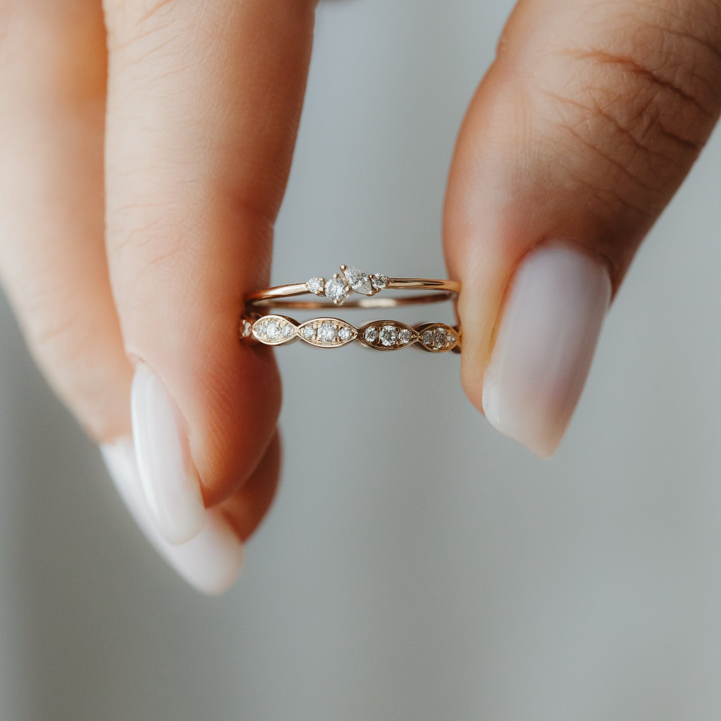Close-up of two 14K yellow gold diamond stacking rings held between the fingers—one with a petite cluster and one with marquise-shaped stations.