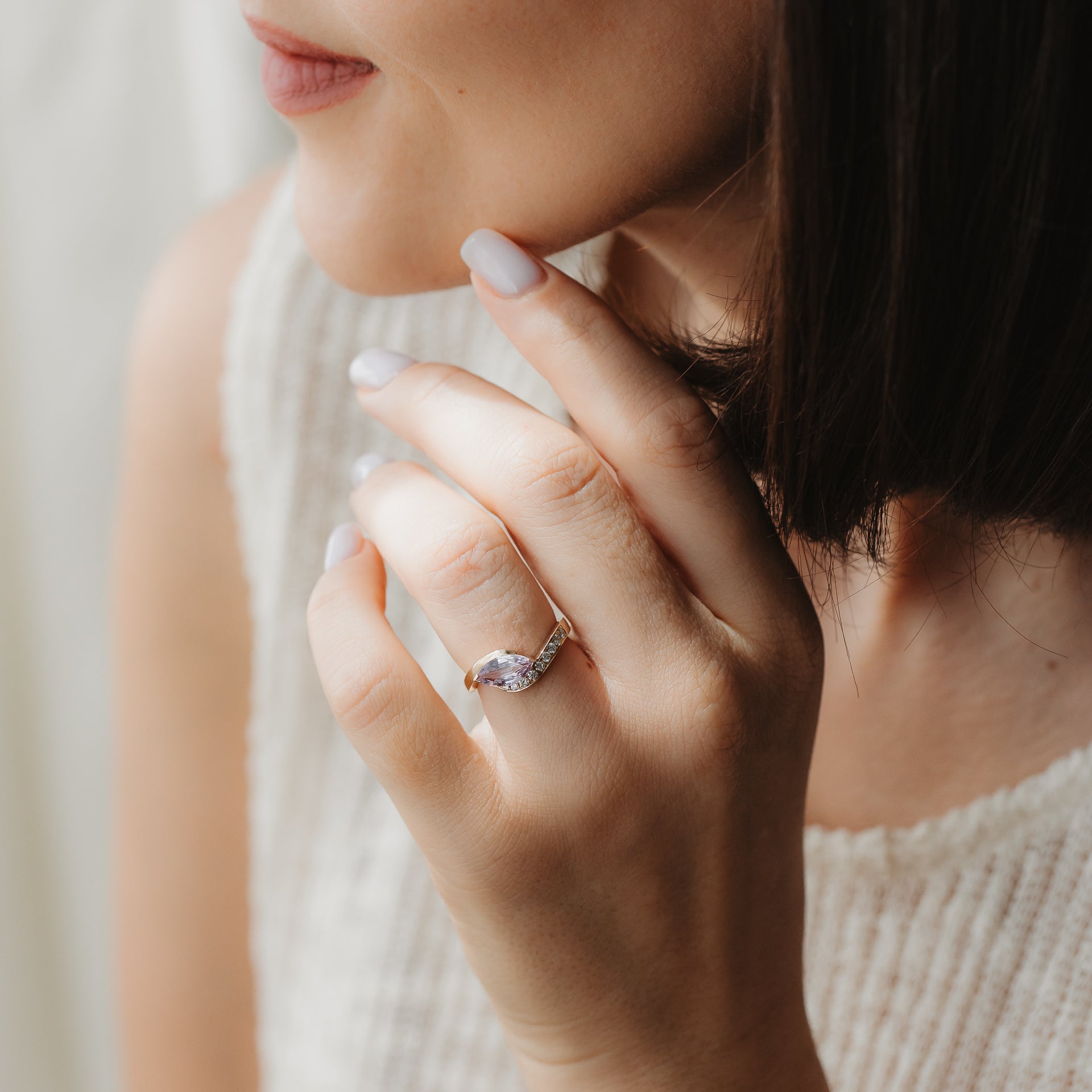 Gros plan d’une main portant une élégante bague de fiançailles en or jaune 14 carats, ornée d’un saphir taille marquise et de diamants, près du visage sous une douce lumière naturelle.
