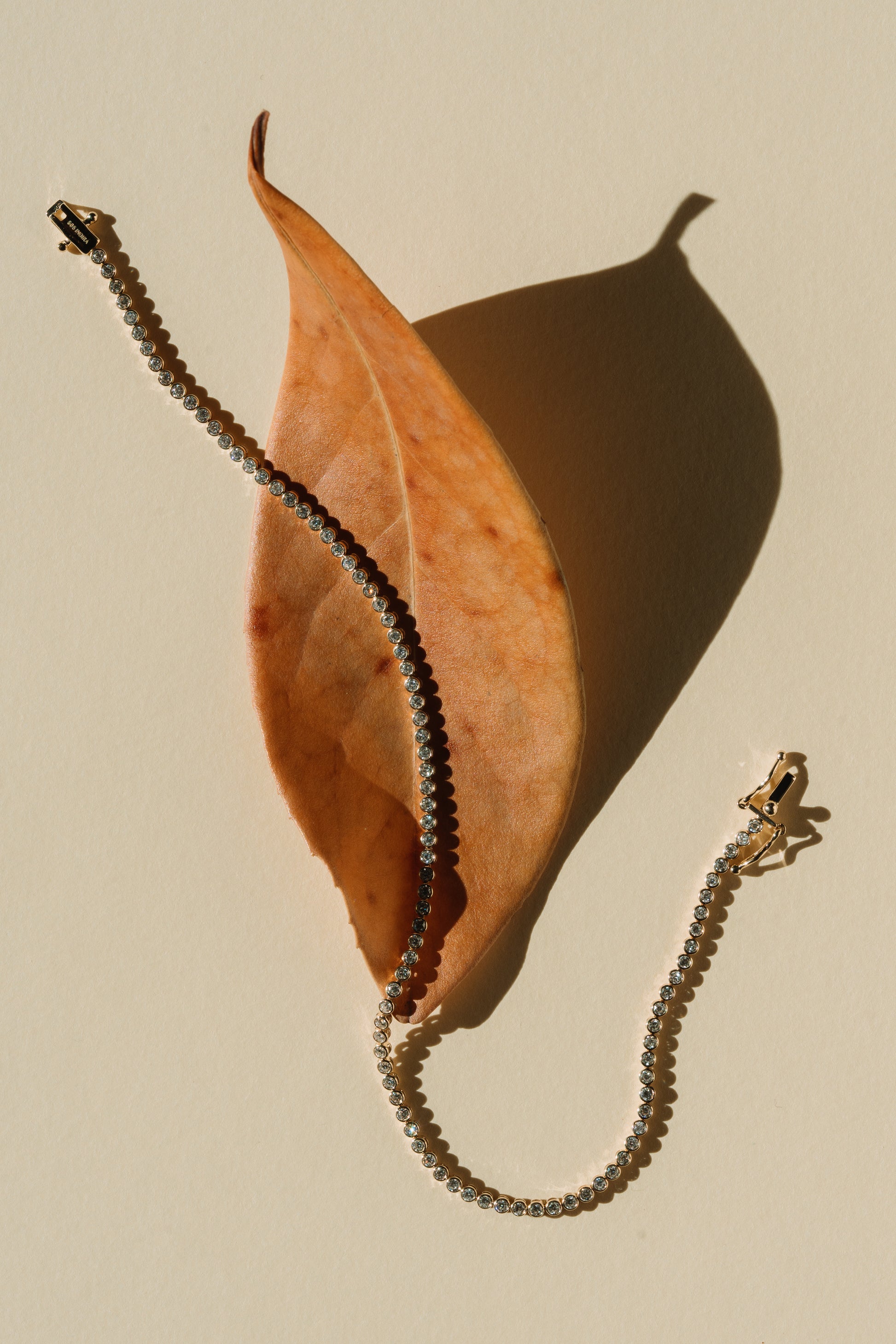 Un élégant bracelet tennis en or serti de diamants, posé sur une feuille d’automne sur un fond beige.