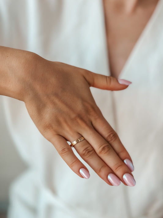 A hand displaying a simple gold wedding band against pink manicured nails.